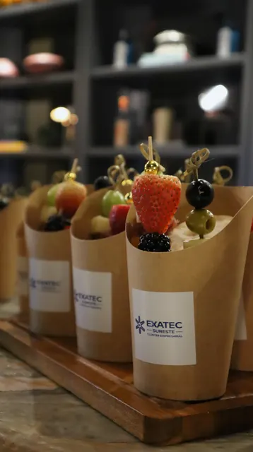 Conos de papel con brochetas de fruta fresca, etiquetados con el logotipo del Clúster Empresarial EXATEC Sureste, sobre una tabla de madera en la recepción.